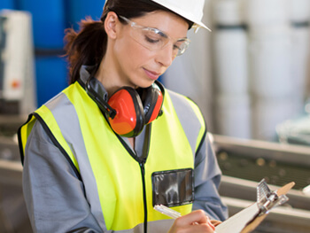 Female technician wearing a safety hat and vest writing on a clip board